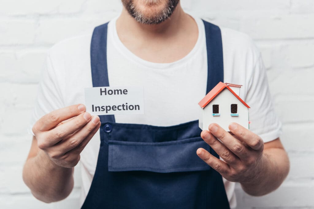 The Importance of Home Inspections in Real Estate.  Male holding a home inspection sign and a small wooden home. 