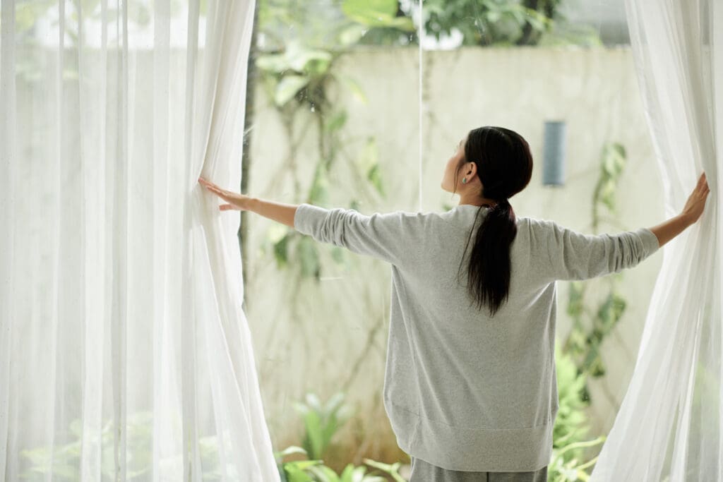 Young woman opening curtains of big window, view from back.  Letting in natural light.